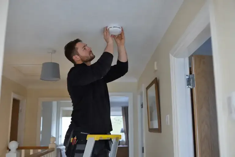 Electrician fitting a smoke alarm to a domestic hallway ceiling.