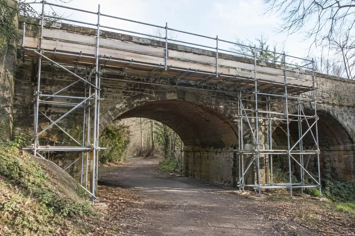 Temporary steel scaffolding providing repair access underneath and along a small British masonry bridge over a dry closed path, multiple braced scaffold bays firmly supported from ground, boarded working platforms with guardrails and toe boards, bridge arches clearly visible, no traffic or workers.