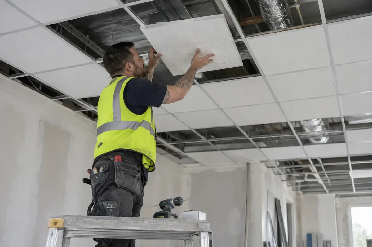 Installer placing an acoustic tile into an exposed suspended ceiling grid.