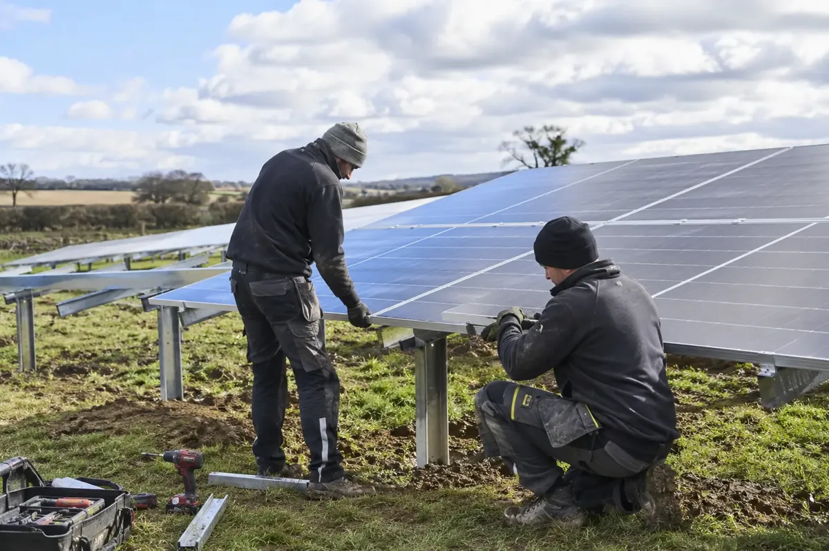 Two installers fastening solar panels onto ground-mounted metal rails in a field.