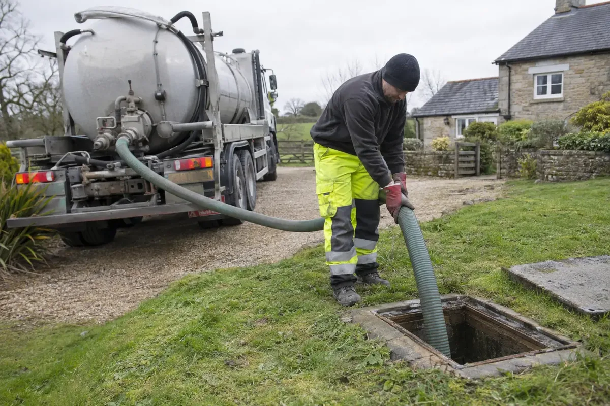 Septic emptying tanker on rural property with large suction hose correctly running to an open septic tank hatch; worker in gloves and protective clothing manages hose from outside the tank, no entry.