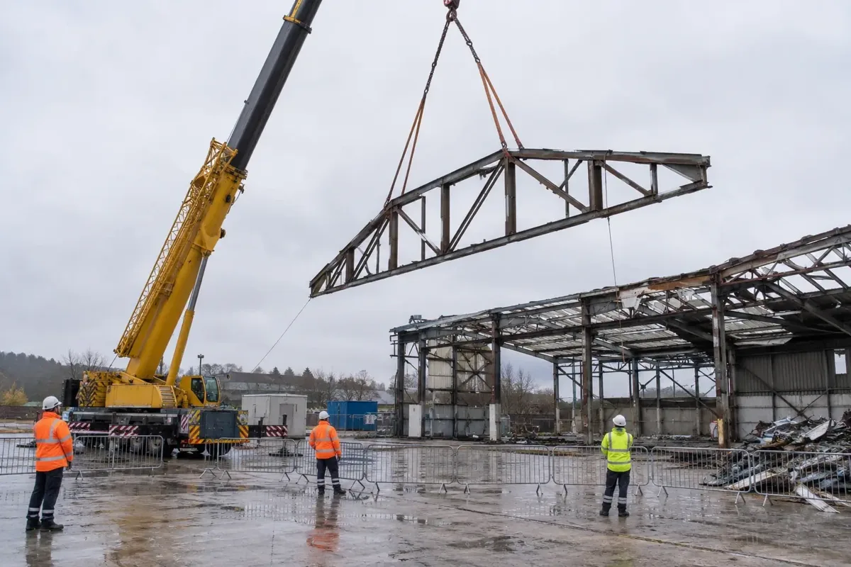 Wide safe view of a crane carefully lifting a detached steel roof truss from a decommissioned industrial building, with a partially dismantled steel frame and controlled exclusion zone. Riggers in PPE stand at safe distance, realistic lifting slings, calm UK industrial site.