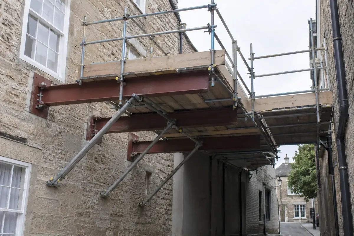 A clearly engineered cantilever scaffold working deck projecting from substantial anchored horizontal steel needle beams through a masonry building facade above a narrow covered passage where ground supports cannot stand; triangulated braces, full guardrails and toe boards, no workers.